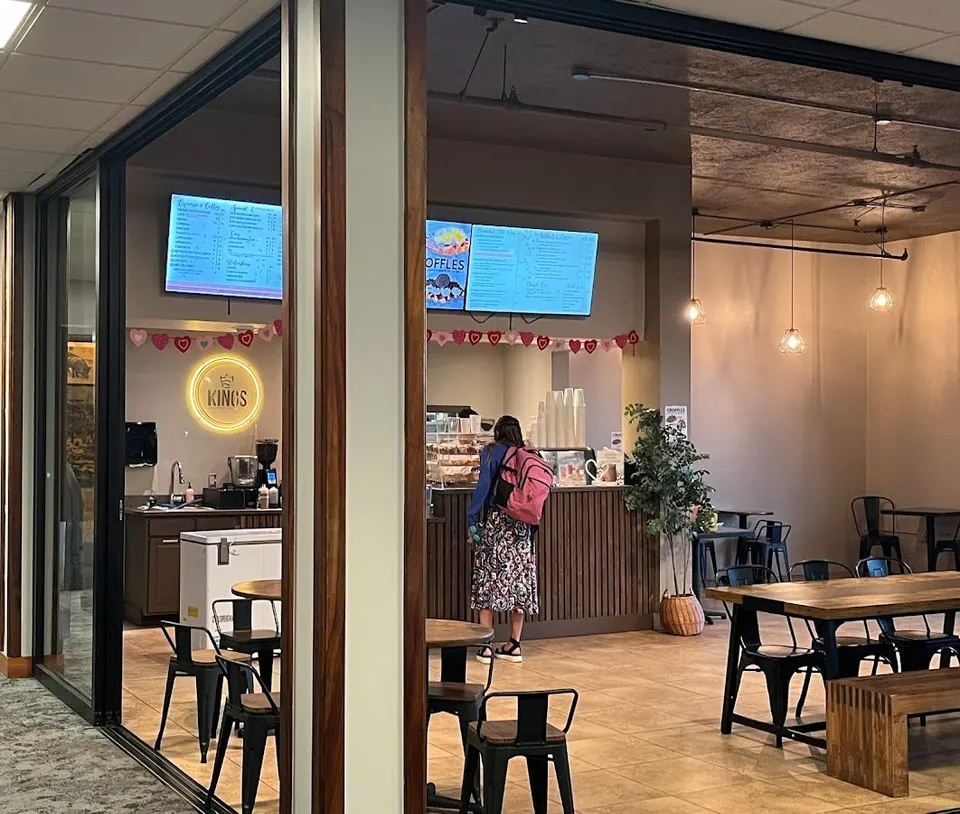 Cafe interior with a lit Kings sign, menu screens, a person at the counter, and tables and chairs.