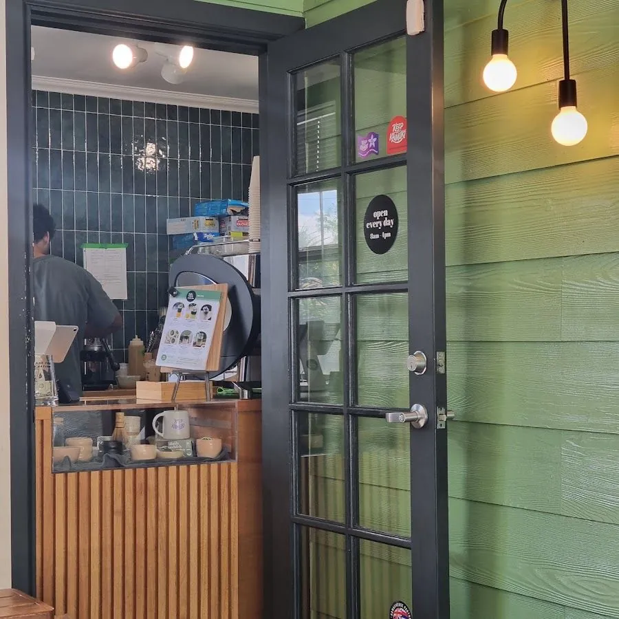 An open black-framed door in a green wall looks into a shop where a worker stands at the counter.