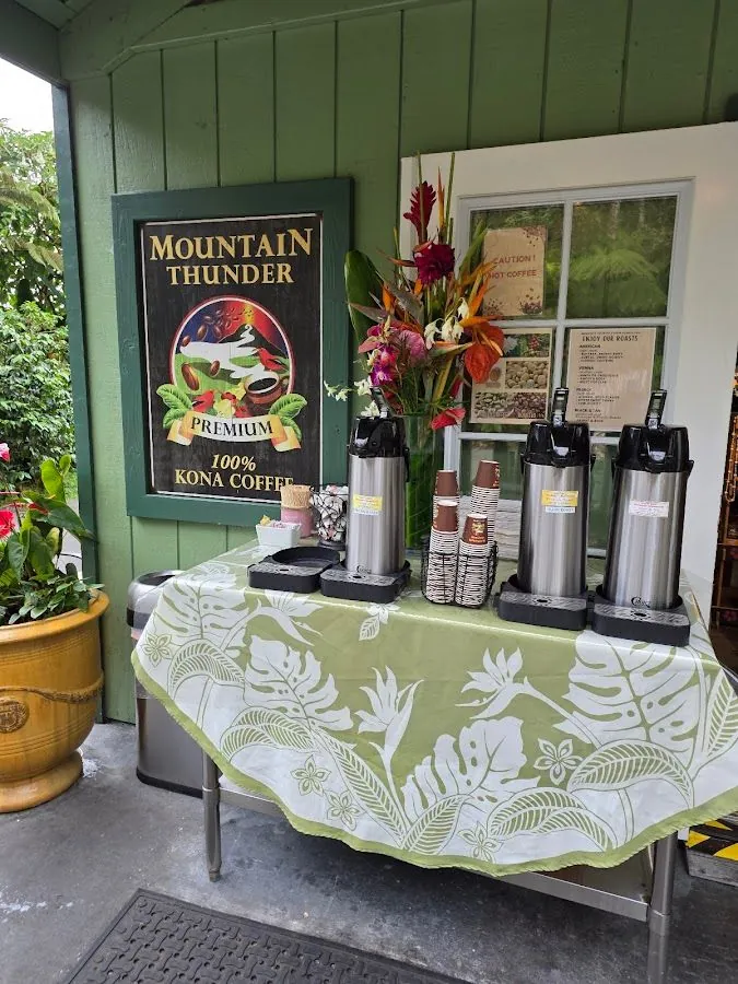 Covered porch with three steel coffee dispensers, cups, tropical flowers, and a Mountain Thunder Kona coffee sign.