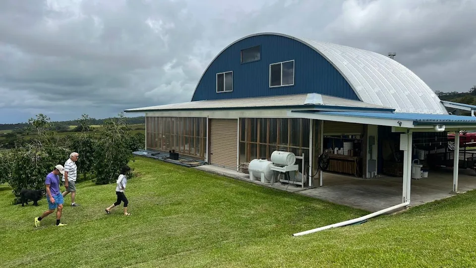 Three people and a black dog walk on grass in front of a blue and silver arched farm building.
