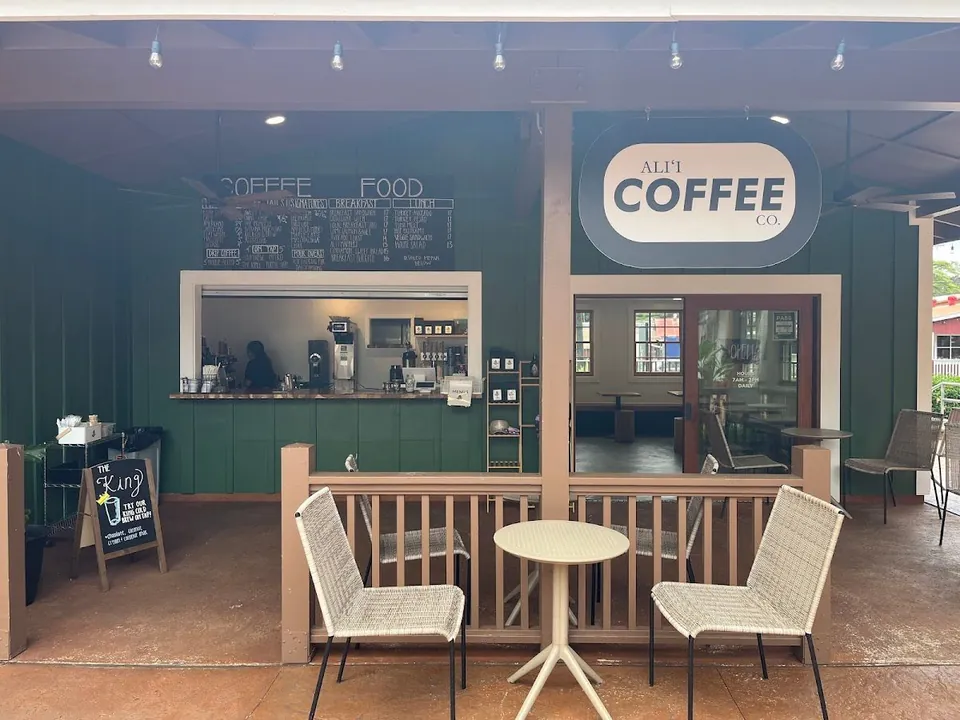 Covered patio and service window of Ali'i Coffee Co., with a wall menu, oval shop sign, and outdoor chairs.