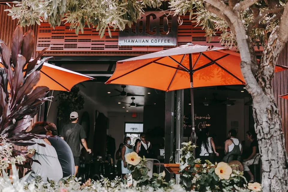 Shop entrance under a Hawaiian Coffee sign, with orange umbrellas, plants, and people seated and standing outside.
