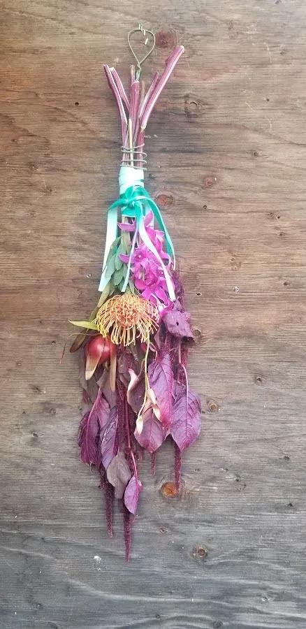 A hook holds wired stems, teal and white ribbons, orange and magenta flowers, and dark purple leaves against wood planks.