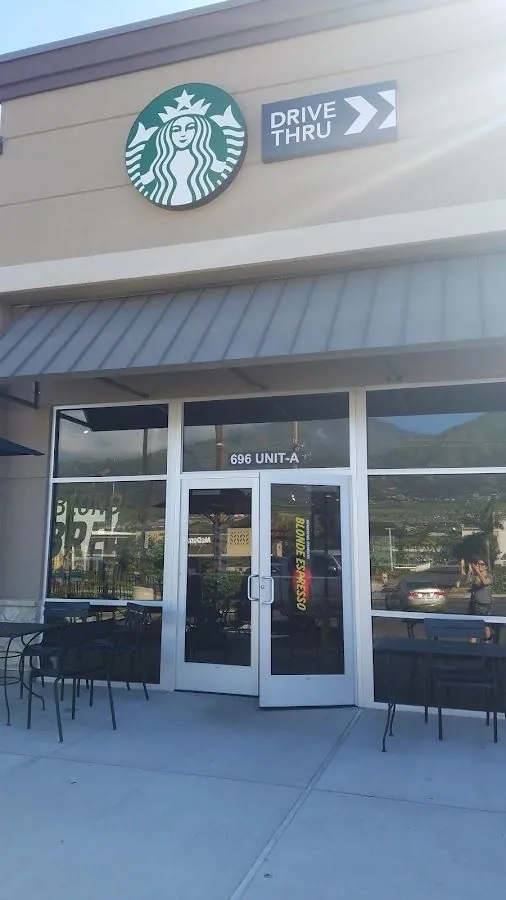 Starbucks storefront with a siren logo, drive-thru sign, glass doors, patio tables, and a person holding a phone.