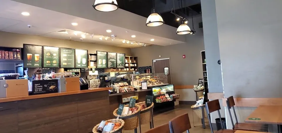 A worker's head shows behind a cafe counter with menu boards, a food case, baskets, and wooden seating.