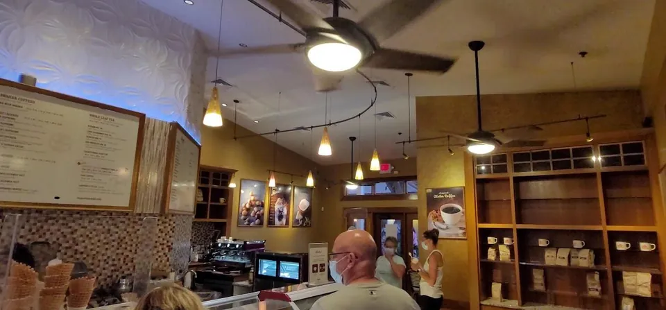 Coffee shop interior with menu boards, service counter, ceiling fans, stocked shelves, and masked customers.