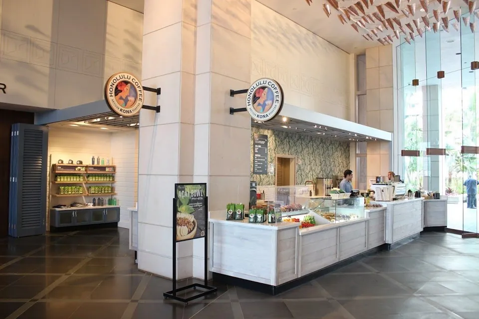 Workers stand behind a stone cafe counter under Honolulu Coffee signs, beside a food case and an acai bowl stand.