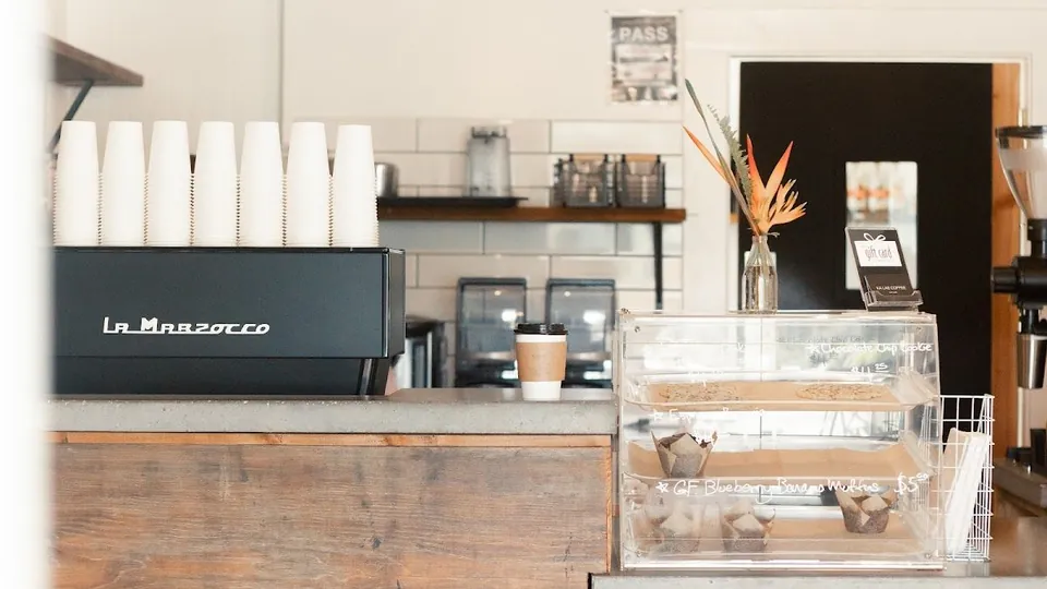 Coffee shop counter with a black espresso machine, stacked cups, a lidded paper cup, and a pastry case.