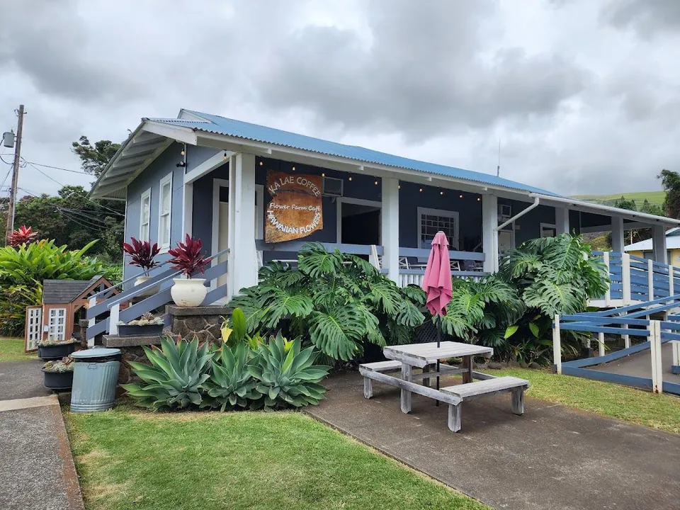 Blue building with a covered porch, wooden Ka Lae Coffee sign, tropical plants, and a picnic table.