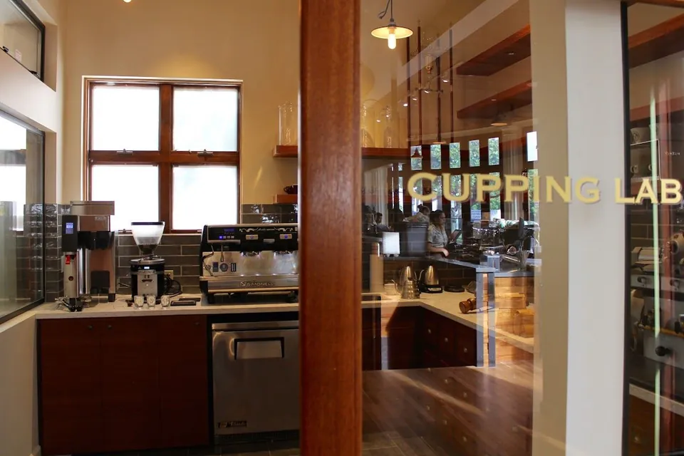 Coffee bar with an espresso machine and grinders beside a glass door labeled CUPPING LAB, where people stand at a counter.