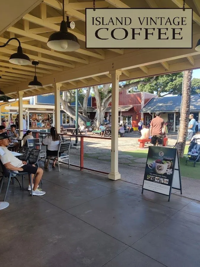 People sit under a hanging Island Vintage Coffee sign beside an A-frame board, with a courtyard beyond the open pavilion.