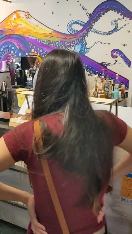 Person with long dark hair stands at a cafe counter beneath an octopus mural, with a coffee grinder nearby.