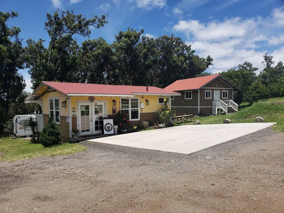 Yellow and brown buildings with red roofs stand by a gravel lot, with a coffee-cup sidewalk sign at the door.