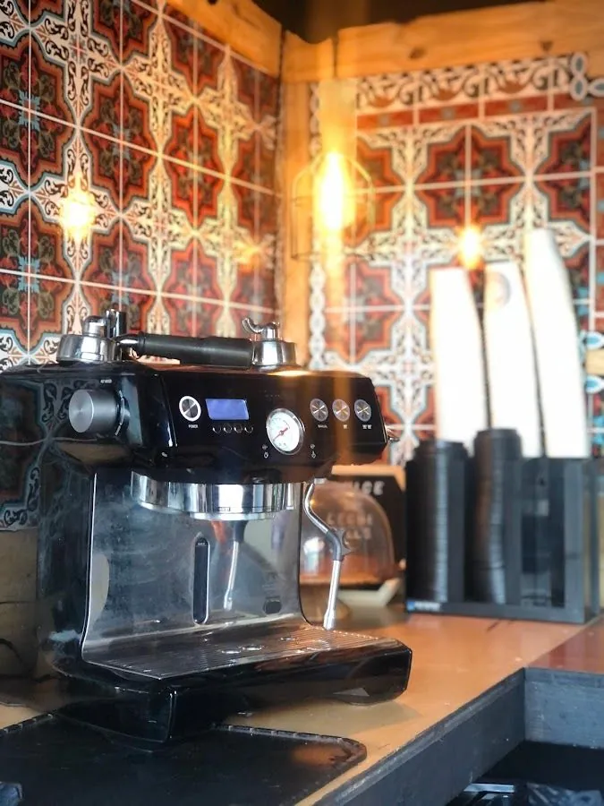 Black espresso machine on a wooden counter, with patterned tile, warm lights, and stacked paper cups.