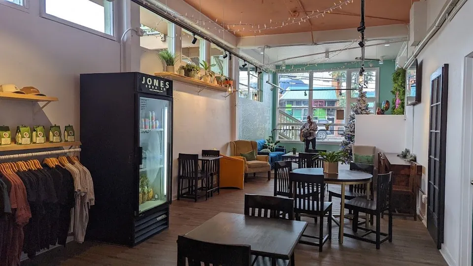 Shop interior with tables, chairs, a Jones Soda cooler, hanging clothes, plants, and string lights.