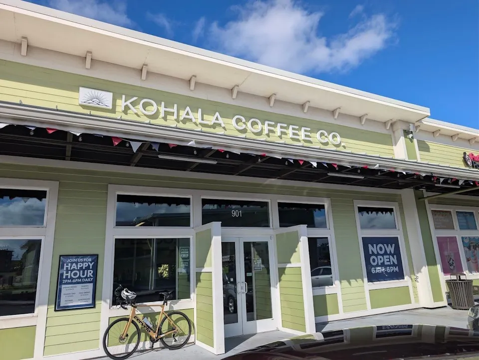 Light green Kohala Coffee Co. storefront with open doors, a parked bicycle, and hours signs under a blue sky.