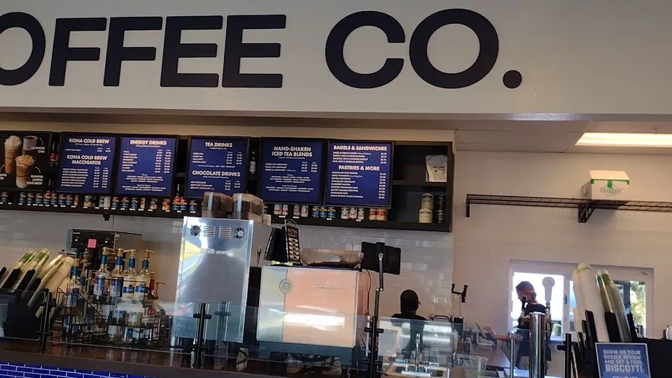 Coffee shop counter with menu boards, espresso machines, cups, and two workers.
