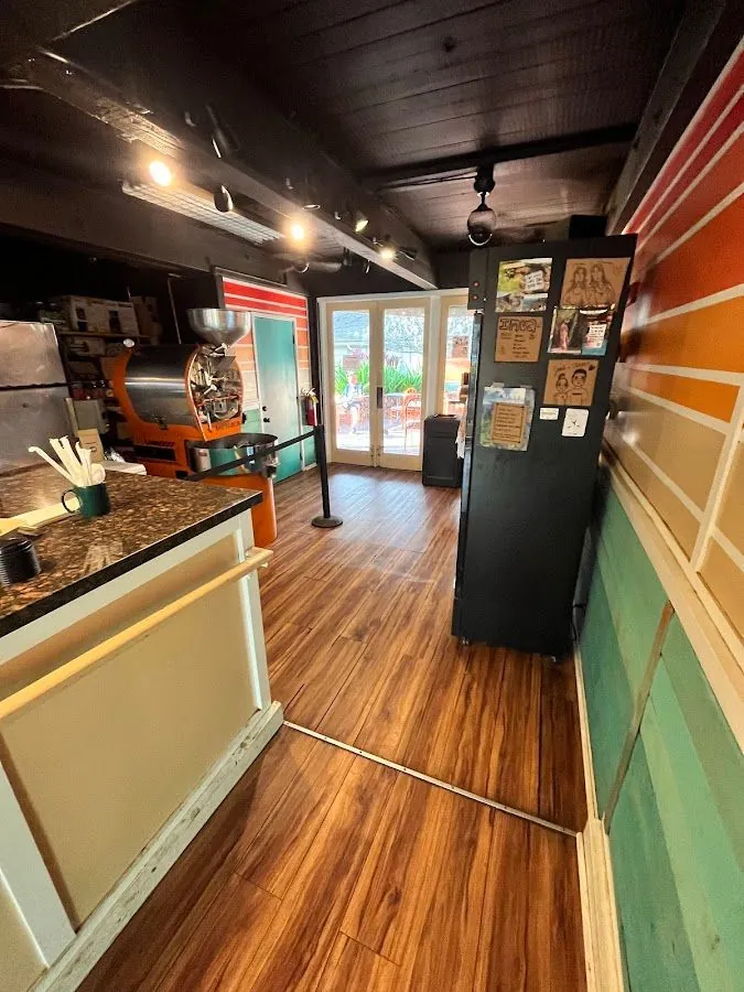 Interior of a coffee roastery with a drum roaster, service counter, wood floors, and glass doors to a patio.