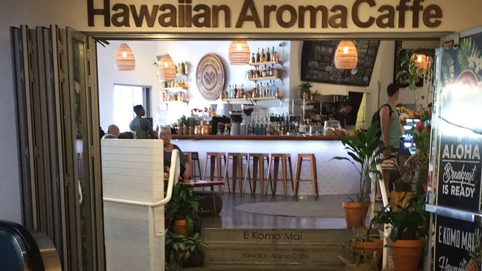 Open entrance of Hawaiian Aroma Caffe showing the counter, wooden stools, hanging lights, plants, and people inside.