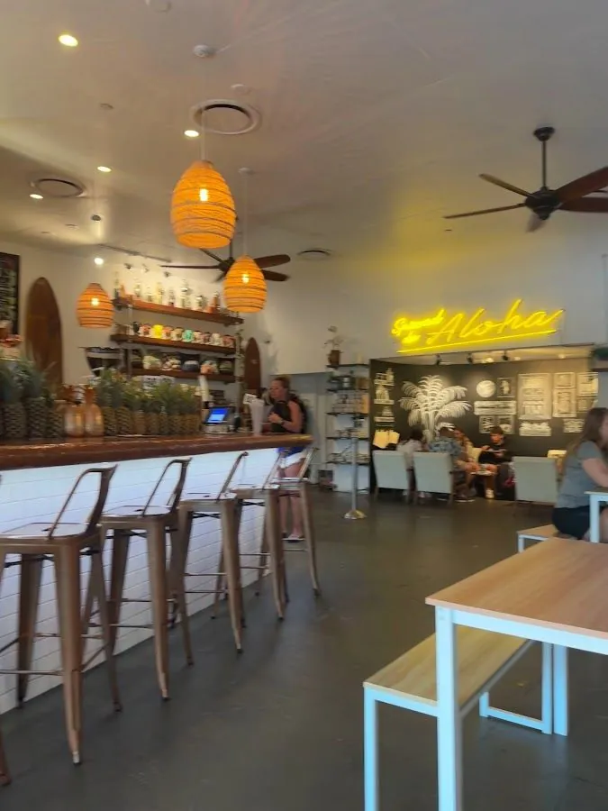 Cafe interior with a wood bar, metal stools, woven pendant lights, and a yellow neon sign reading Spread the Aloha.