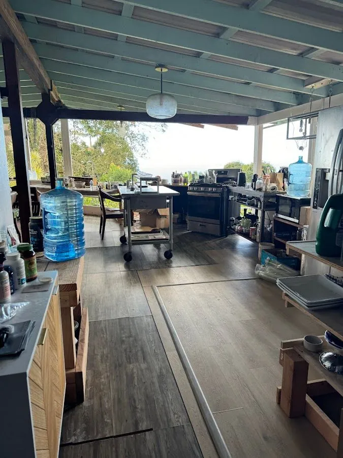 Open kitchen under a painted wood roof, with a stove, a cart, water jugs, and trees outside.