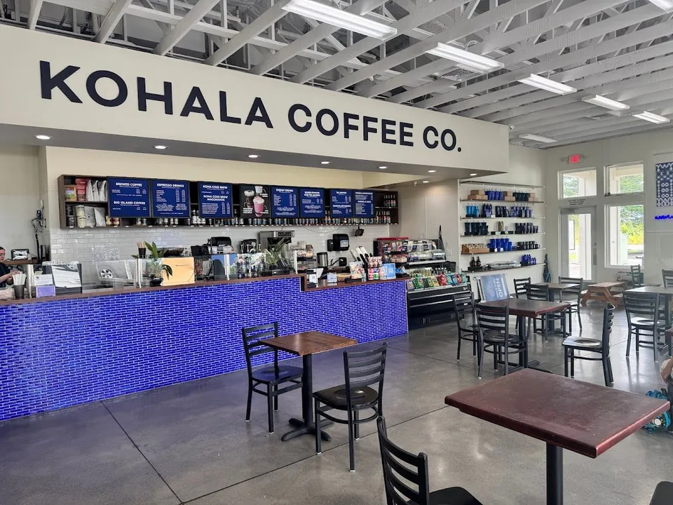 Cafe interior with a large Kohala Coffee Co. sign, blue-tiled service counter, menu boards, tables, and retail shelves.
