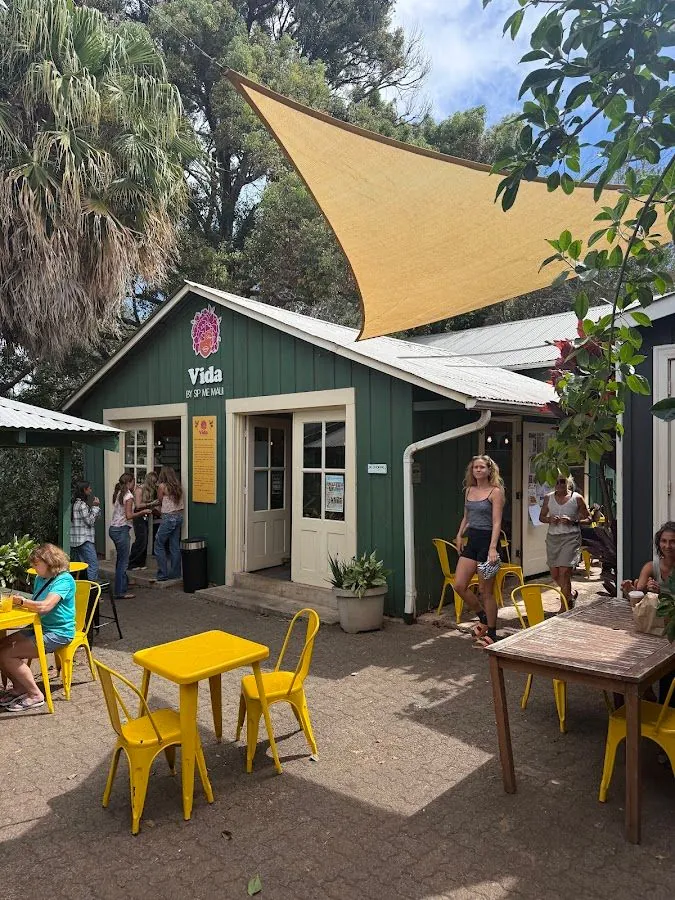 People sit and stand outside the green Vida by Sip Me Maui shop under a shade sail.