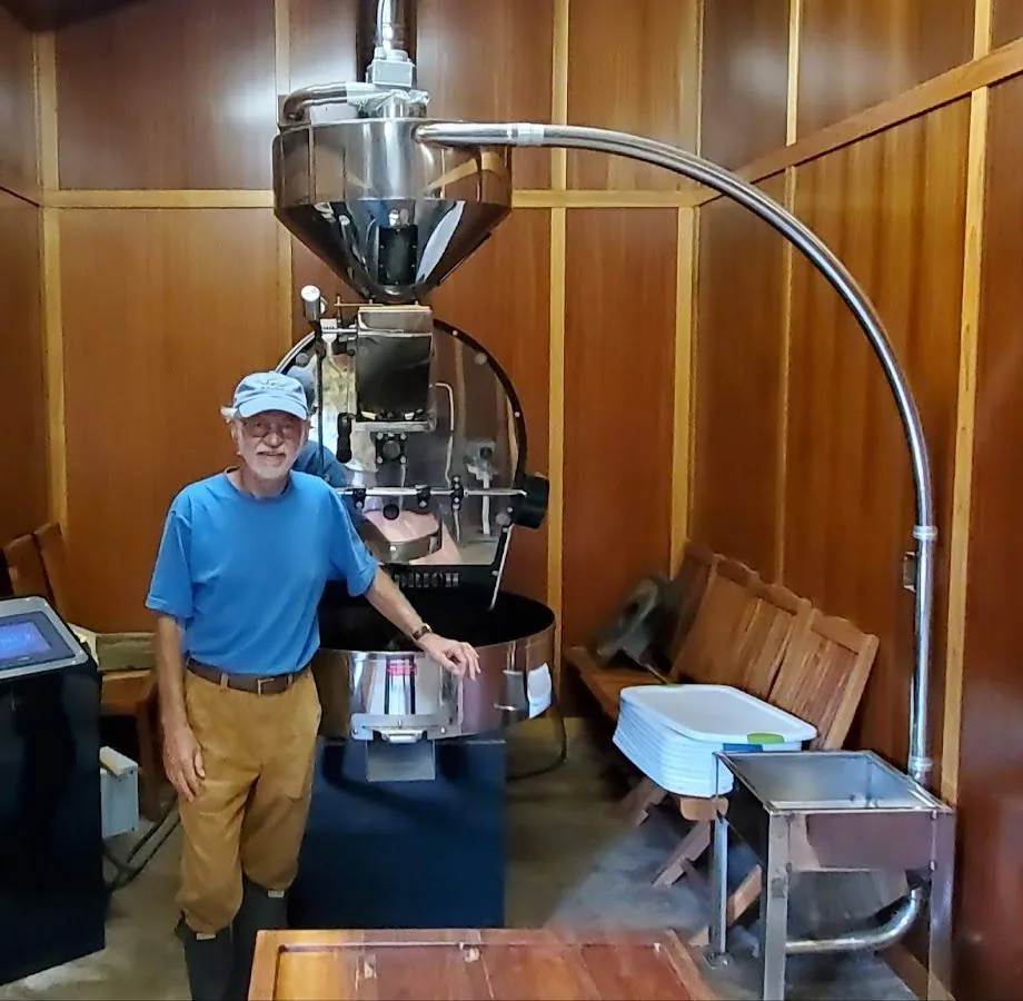 A person in a blue shirt and cap stands beside a large stainless steel coffee roaster in a wood-paneled room.