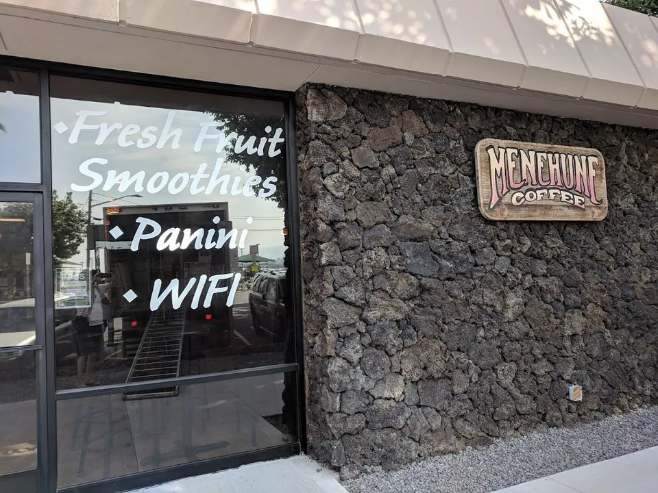 Lava-rock storefront of Menehune Coffee with a wooden sign and window text for smoothies, panini, and WiFi.