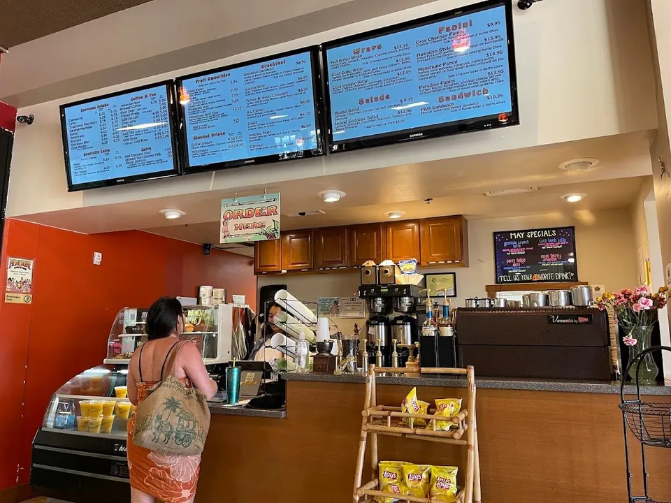 Cafe service counter with overhead menu screens, an order sign, espresso equipment, and two people at the counter.