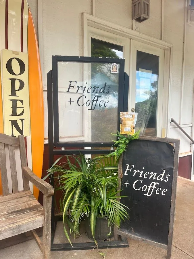 Shop entrance with Friends + Coffee signs, an OPEN board, a surfboard, a wooden bench, and potted plants.