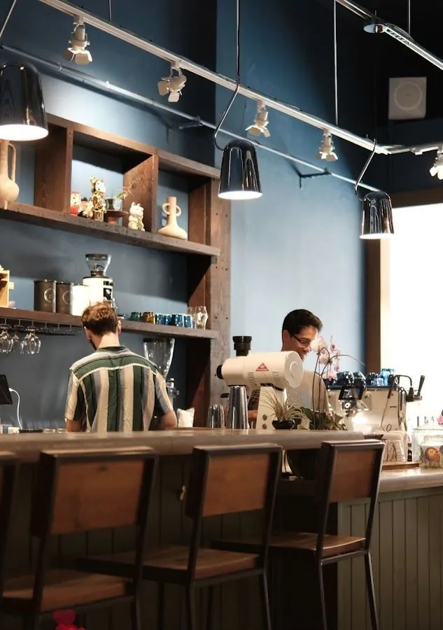Two workers stand behind a wooden cafe counter with stools, grinders, shelves, and pendant lights.