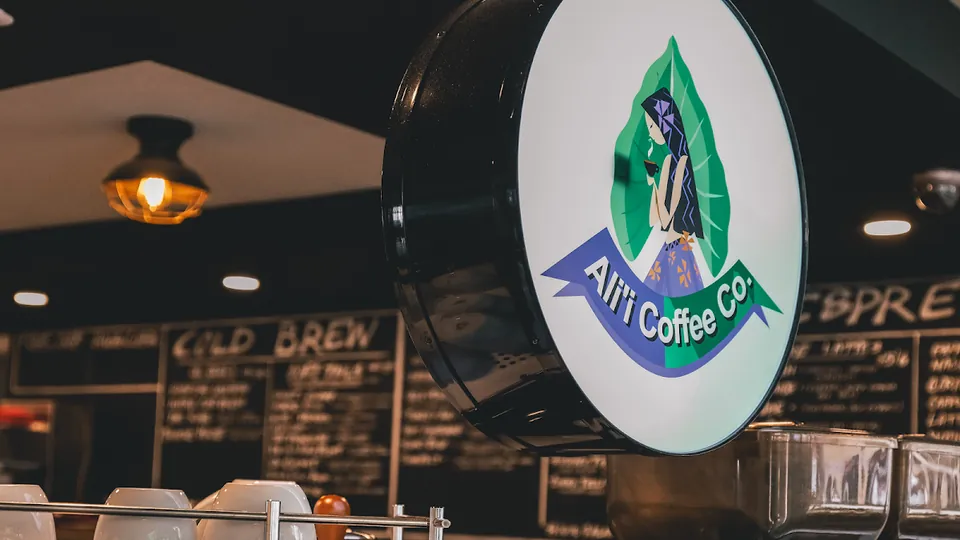 A round Ali'i Coffee Co. sign hangs in a shop interior above cups, with chalkboard menus behind it.