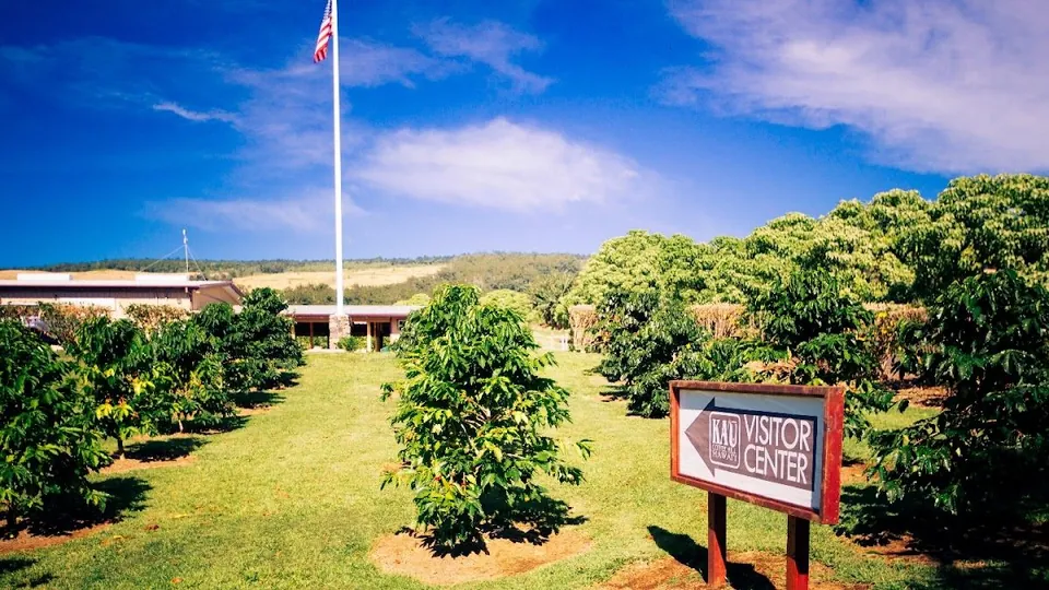 A visitor center sign stands on a lawn of coffee trees, with a US flag and a low building behind it.