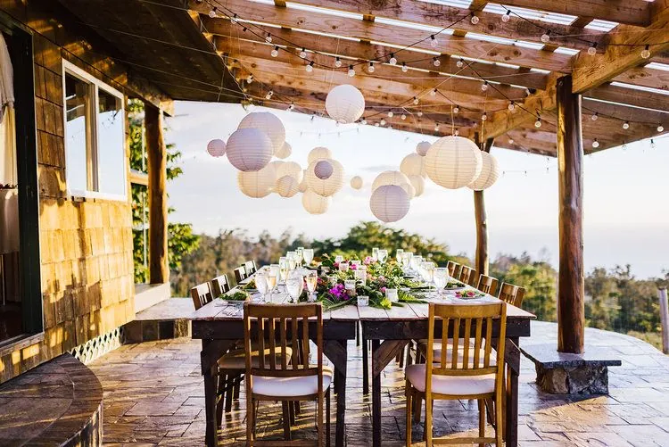 A long table set with glassware and flowers sits under a wooden pergola hung with lanterns and string lights.