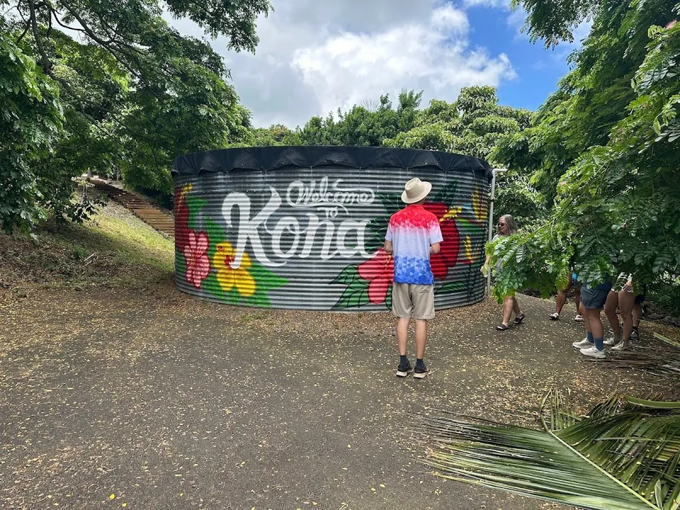 A person in a hat faces a flower-painted tank reading Welcome to Kona while others walk under nearby trees.