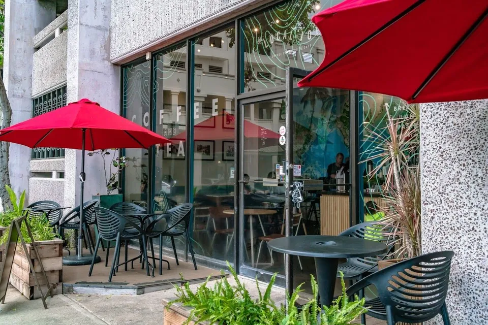 Storefront of a coffee shop with red patio umbrellas, black outdoor chairs and tables, glass windows, and plants.