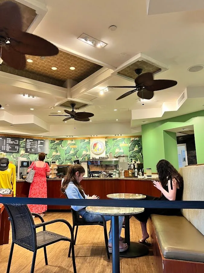 People sit with phones and stand at a cafe counter beneath ceiling fans and a leaf-pattern wall.
