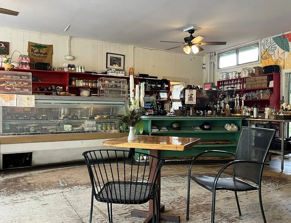 A coffee shop interior shows seating, a glass display case, red shelves, and a person behind the counter.