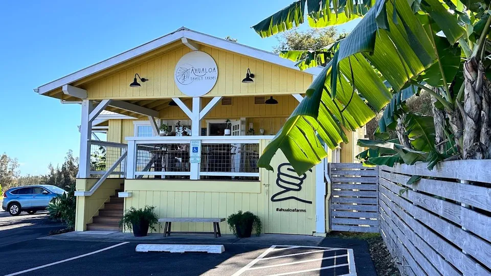 Yellow gabled storefront signed Ahualoa Family Farms, with a porch, bench, and banana leaves.