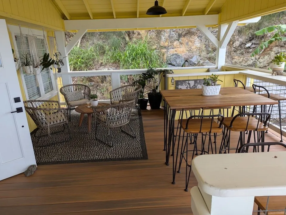 Yellow covered porch with woven chairs, a wood table, and stools open to a rocky green hillside.