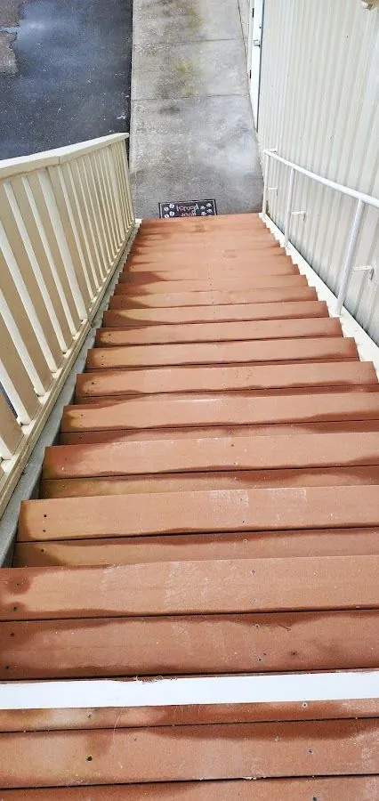 Reddish-brown outdoor stairs with pale railings descend to a concrete walkway beside a metal wall.