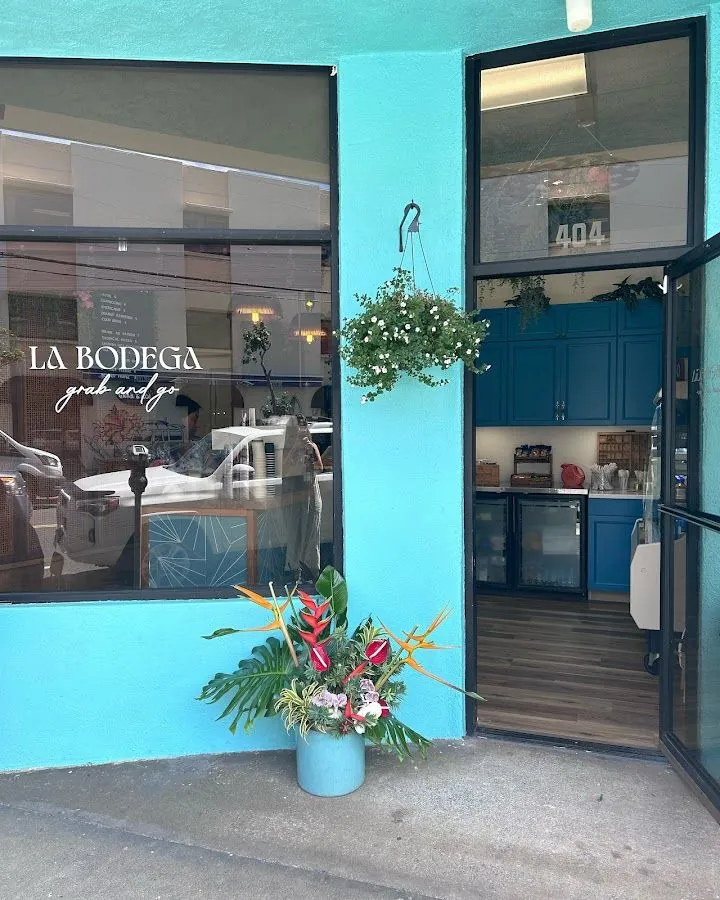 Turquoise storefront labeled LA BODEGA grab and go, with an open door, hanging plant, and sidewalk flower pot.