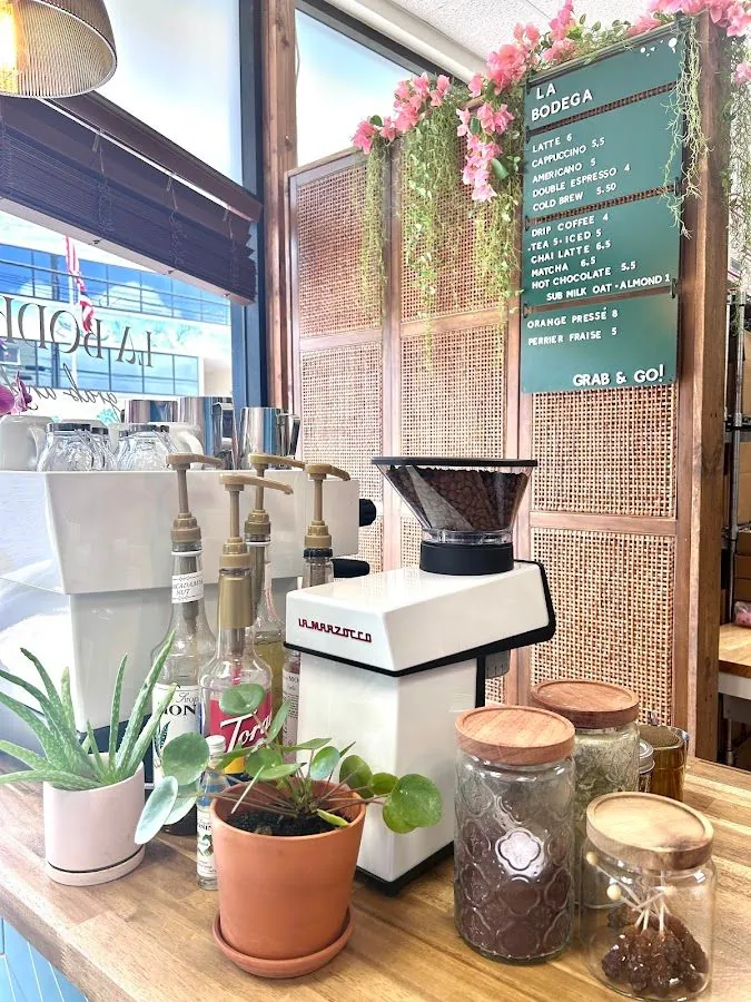 Coffee counter with a white grinder, plants, glass jars, and a green La Bodega menu board.