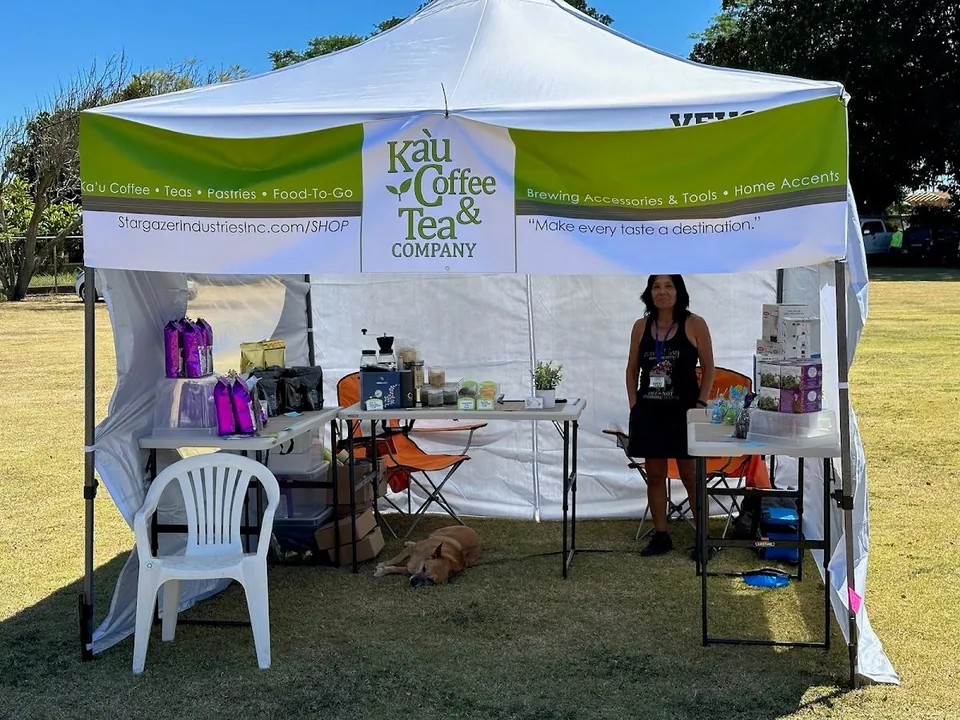 Ka'u Coffee Tea and Company canopy booth on a grassy field, with product tables, a standing person, and a dog.