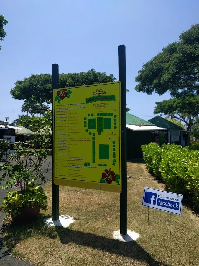 Yellow outdoor directory sign for Ali'i Gardens Marketplace, with plants and a Facebook yard sign.