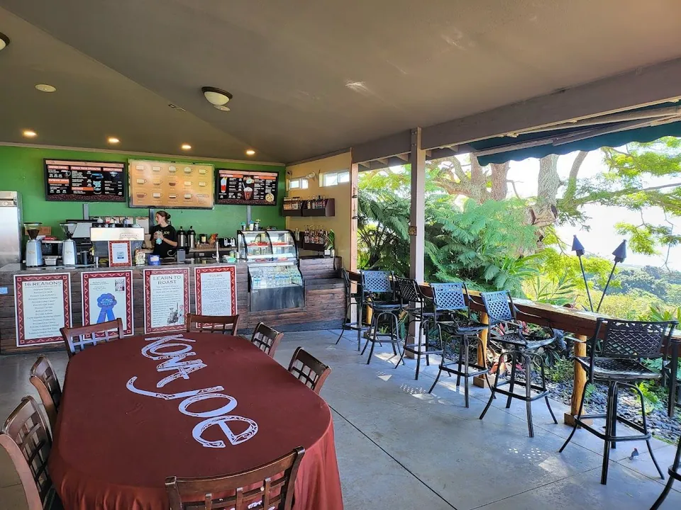 Covered cafe counter with menu boards, a Kona Joe tablecloth, bar stools, and a tropical view.