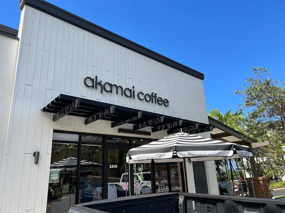 White storefront signed akamai coffee, with striped patio umbrellas and outdoor seating under a clear blue sky.