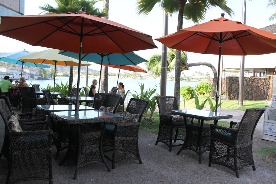 People sit at wicker patio tables under orange umbrellas beside palm trees and water.
