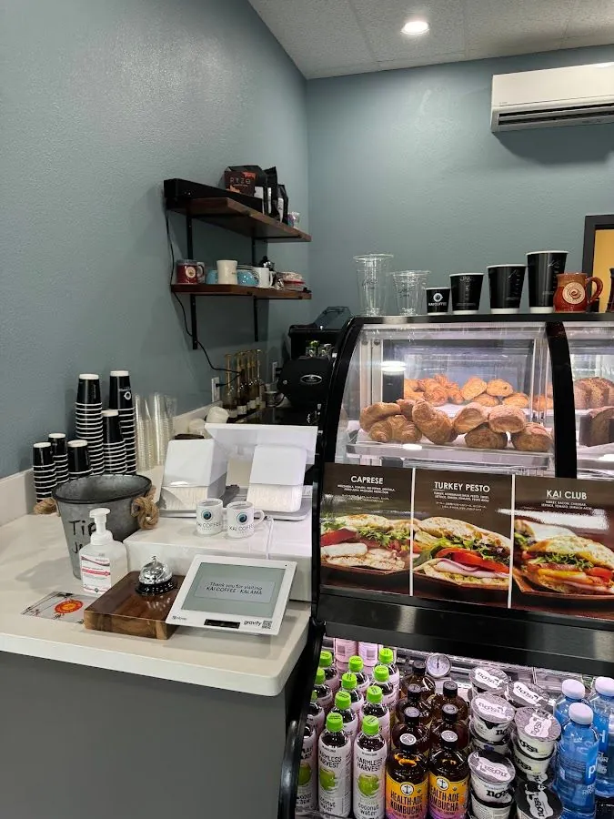 Cafe counter with pastry case, sandwich cards, bottled drinks, and a Kai Coffee Kalama welcome screen.